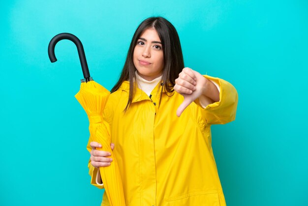 写真 防雨コートと青の背景に分離された傘を持つ若い白人女性が否定的な表現で親指を下に示す