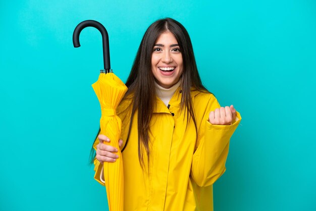 Young caucasian woman with rainproof coat and umbrella isolated on blue background celebrating a victory in winner position