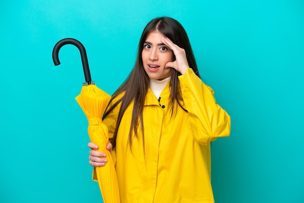 Giovane donna caucasica con cappotto antipioggia e ombrello isolato su sfondo blu che fa un gesto a sorpresa mentre guarda di lato