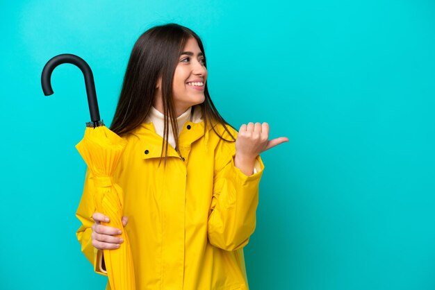 Young caucasian woman with rainproof coat and umbrella isolated on blue background pointing to the side to present a product