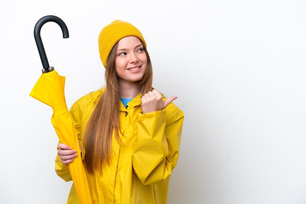 Young caucasian woman with rainproof coat and umbrella isolated on white background pointing to the side to present a product