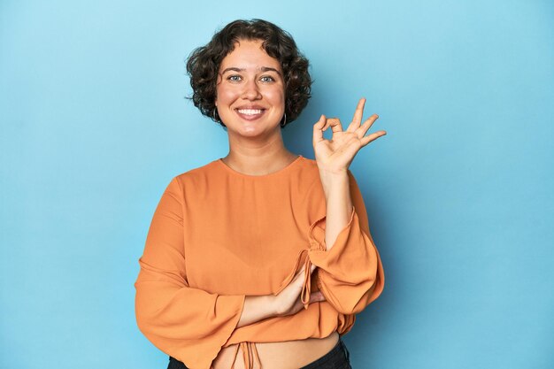 Young caucasian woman with short hair winks an eye and holds an okay gesture with hand