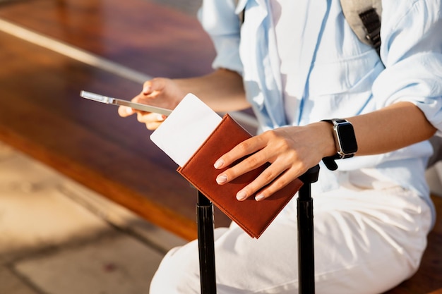 Young caucasian woman with suitcase passport and ticket typing on smartphone waiting for transport