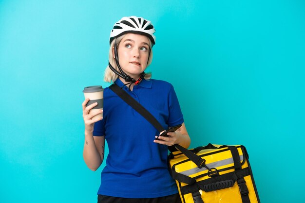 Young caucasian woman with thermal backpack isolated on blue background holding coffee to take away and a mobile while thinking something