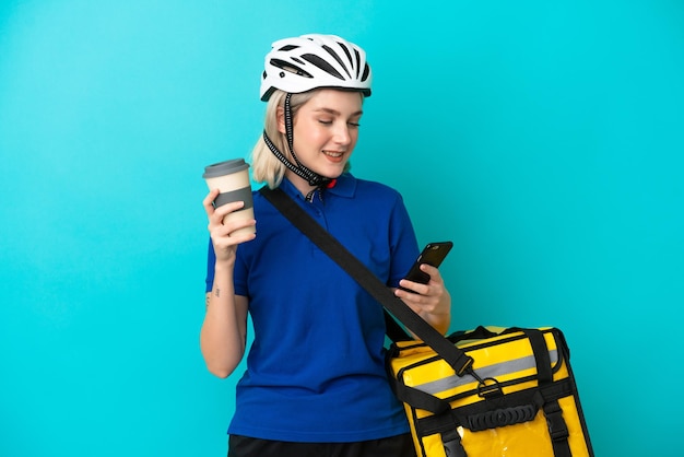 Young caucasian woman with thermal backpack isolated on blue background holding coffee to take away and a mobile