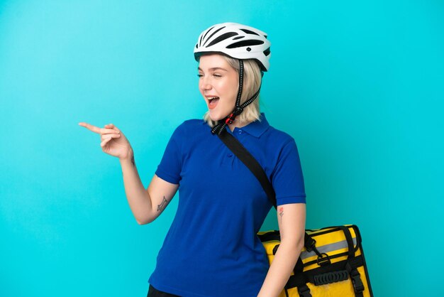 Young caucasian woman with thermal backpack isolated on blue background pointing finger to the side and presenting a product