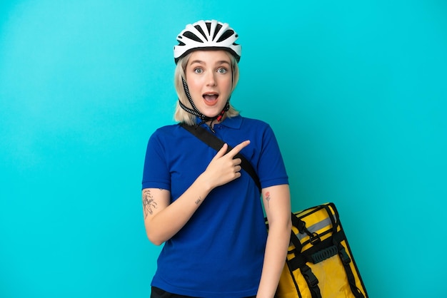 Young caucasian woman with thermal backpack isolated on blue background surprised and pointing side