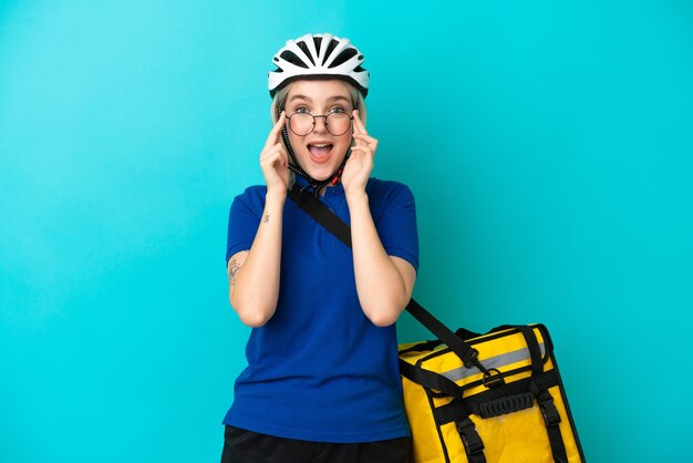 Young caucasian woman with thermal backpack isolated on blue background with glasses and surprised