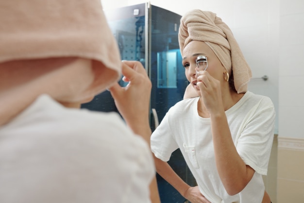 Young Caucasian woman with towel on head using eyelash curler while standing in front of mirror in bathroom