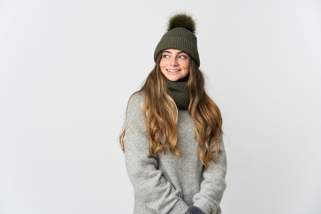 Young caucasian woman with winter hat isolated on white wall looking to the side and smiling