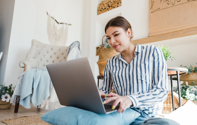 Young caucasian woman working on laptop in cozy living room, sitting on the floor with pillows.