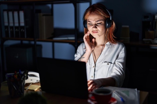 Young caucasian woman working at the office at night touching mouth with hand with painful expression because of toothache or dental illness on teeth dentist