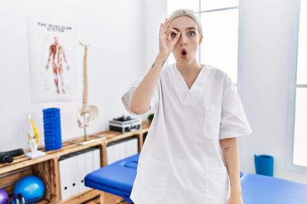 Молодая кавказская женщина, работающая в клинике по восстановлению боли, делает нормальный жест, потрясенная удивленным лицом, глядя сквозь пальцы. недоверчивое выражение.