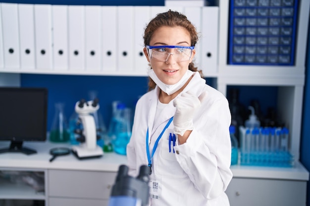 Young caucasian woman working at scientist laboratory pointing thumb up to the side smiling happy with open mouth