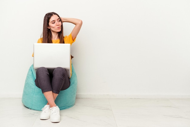 Young caucasian woman working with laptop sitting on a puff isolated on white background touching back of head, thinking and making a choice.