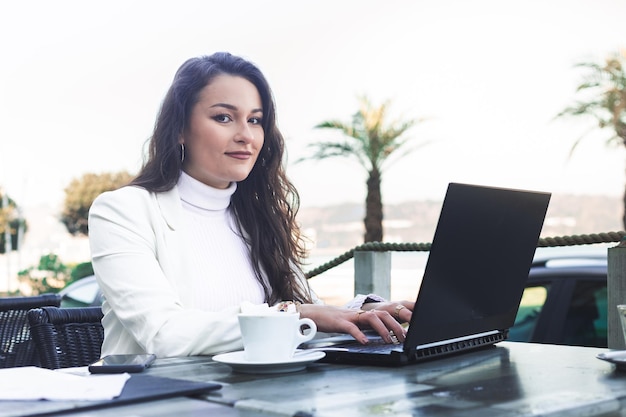 Young caucasian woman working with a laptop while taking a coffee break at a terrace next to the beach