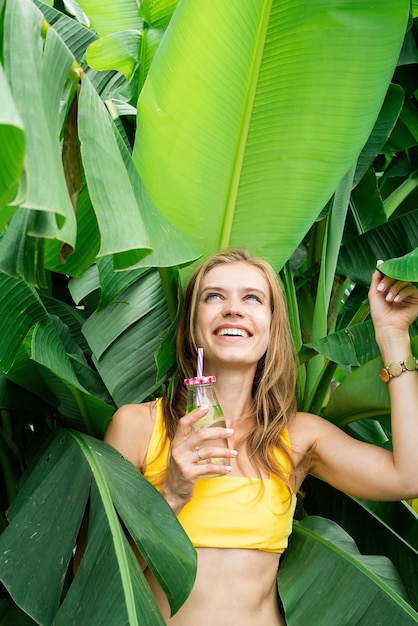 Young caucasian woman in yellow swuimsuit drinking beverage standing in tropical palm leaves