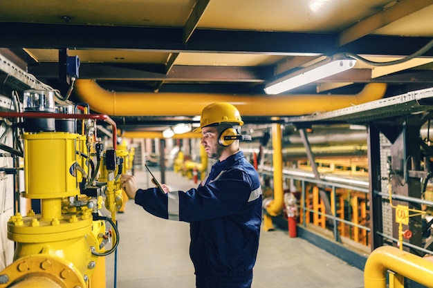 Giovane lavoratore caucasico in vestito protettivo facendo uso della compressa mentre controllando le macchine nell'impianto di riscaldamento.