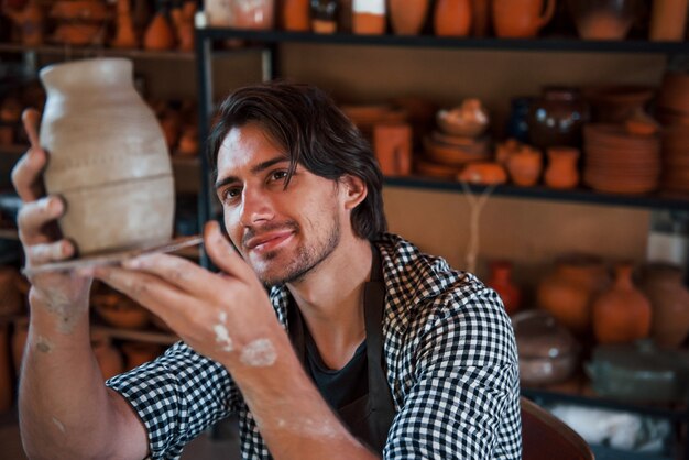 Young ceramist holds fresh handmade pot in hand and looks at results of his work.