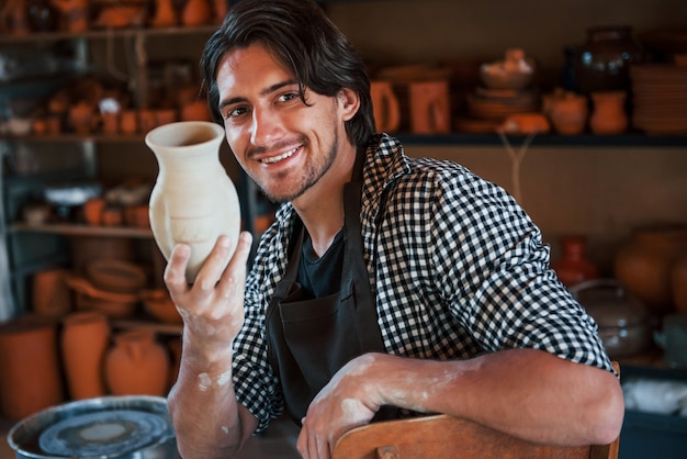 Фото Молодой керамист держит в руке свежий горшок ручной работы. доволен результатами своей работы.