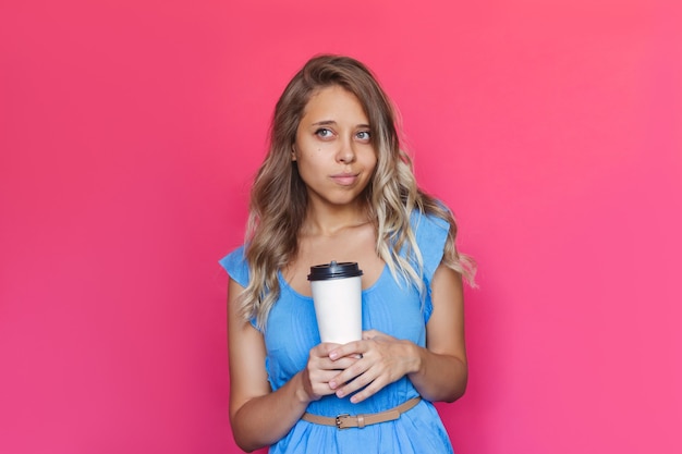 A young charming blonde woman holds a white paper cup of coffee or tea with copy space looking away