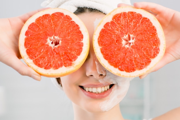 Young charming girl holding two halfs of grapefruit in hands, covering her eyes. Healthy diet food. Natural cosmetics, skincare, wellness, facial treatment, cosmetology concept.