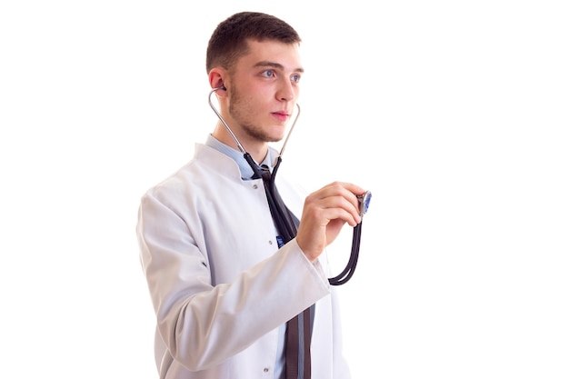 Young charming man with brown hair in blue shirt tie and doctor gown using stethoscope in studio