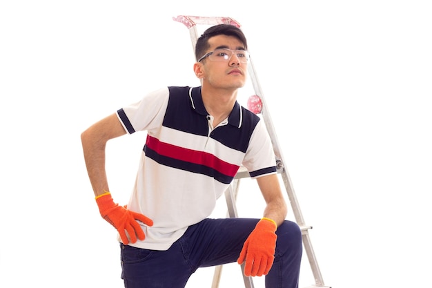 Young charming man with dark hair in white and blue tshirt and jeans with orange gloves and protective glasses standing near the ledder with electric screwdriver on white background in studio