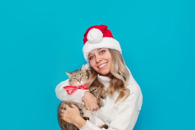 A young charming smiling blonde woman in a white warm sweater, Santa hat holds adorable Tabby cat