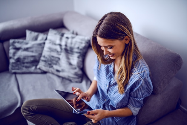 Young charming smiling caucasian brunette in stripped shirt sitting on sofa in living room and using tablet.