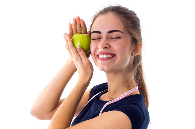 Young charming woman in blue T-shirt with centimeter around her neck holding green apple near the face on white background in studio