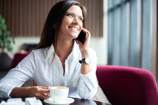 自由時間にコーヒーショップで一人で座ってスマートフォンで呼び出す若い魅力的な女性