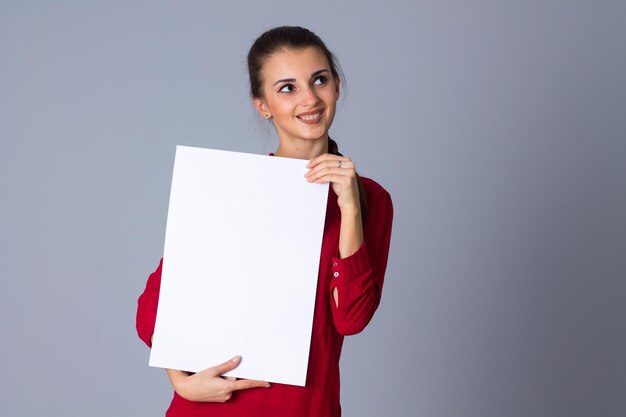 Young charming woman holding white sheet of paper and thinking on grey background in studio