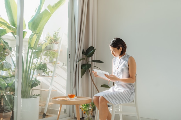 Photo young charming woman sitting on chair and reading book next to window at home.