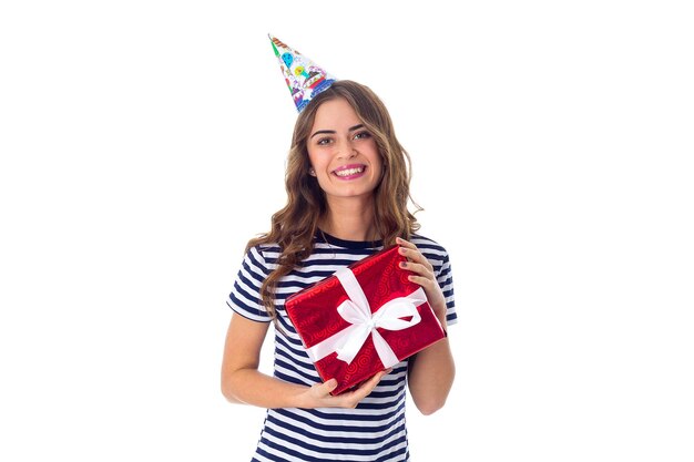 Young charming woman in stripped T-shirt and celebration cap with long hair holding red present on white background in studio