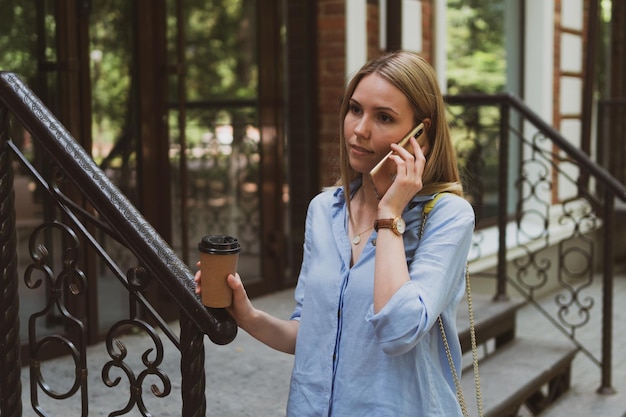 写真 街の通りで電話で話している彼女の手にコーヒーを持つ若い魅力的な女性