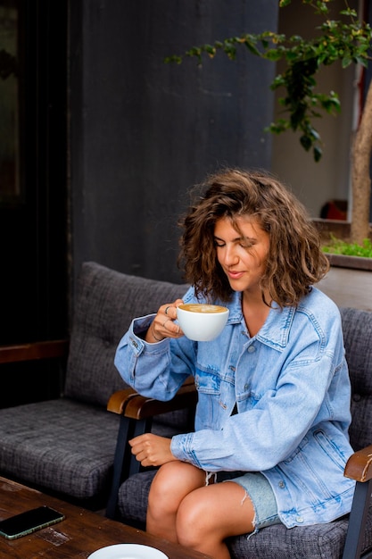 Young charming woman with curly hairstyle holding big mug having fresh cappuccino