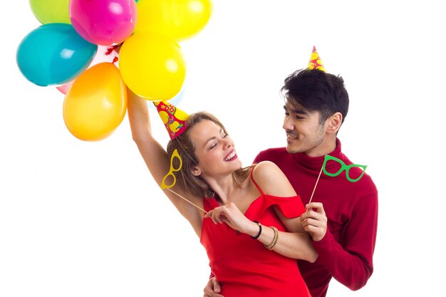 Young charming woman with long chestnut hair in red dress and young handsome man in red shirt with celebrating hats holding many colored balloons and card sticks of glasses on white background in stud