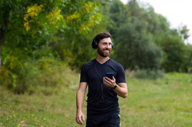 Photo young cheerful active guy sporty walking with headphones mobile phone enjoying listening to music, jogging outdoors on urban city park or forest background.