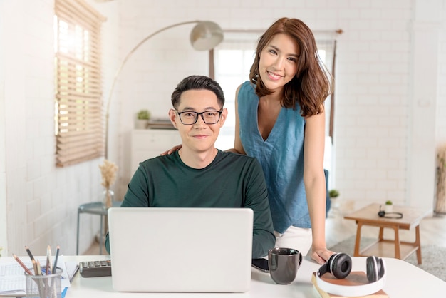 若い陽気なアジアのカップルの同僚は、お互いに話しているオフィスの屋内でラップトップコンピューターで働いています。