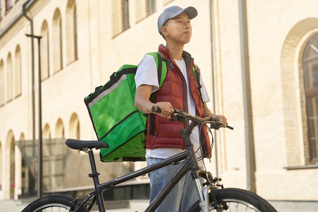 日当たりの良い通りに彼の自転車で立っているサーモバッグを持つ若い陽気なアジア人男性の宅配便。フードデリバリーコンセプト