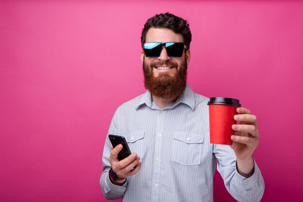 Young cheerful bearded man holding a take away cup and his phone on pink background.