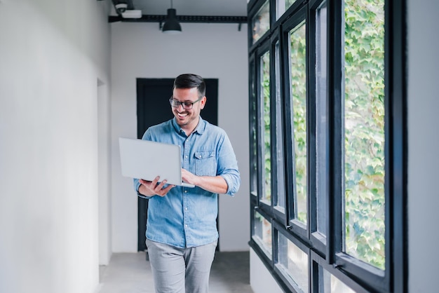 Young cheerful beautiful joyful man manager businessman entrepreneur small business owner employee worker with glasses smiling while working with his laptop walking in office hallway corridor