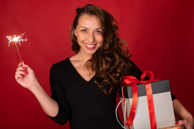 Young cheerful beautiful woman with gift box and burning sparkler smiling looking at camera on red background. Christmas holiday party, New Year's Eve celebration, gifting ideas concept. Copy space