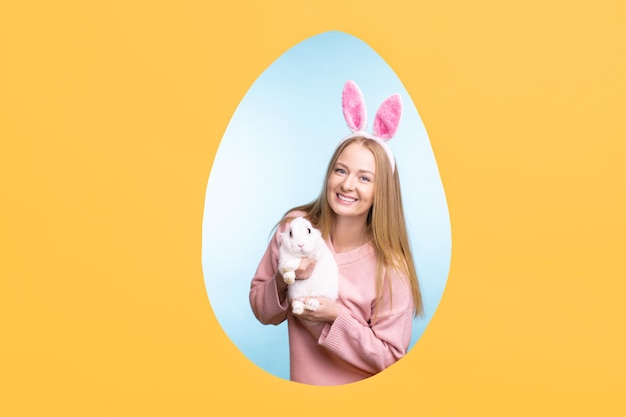 Young cheerful blond woman in bunny headband holding fluffy easter rabbit