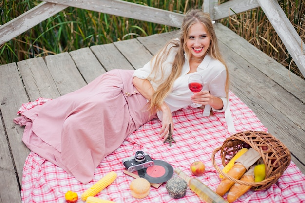 ヴィンテージの服の若い陽気なブロンドの女性は木製の桟橋でピクニックテーブルクロスにある間赤ワインを飲む