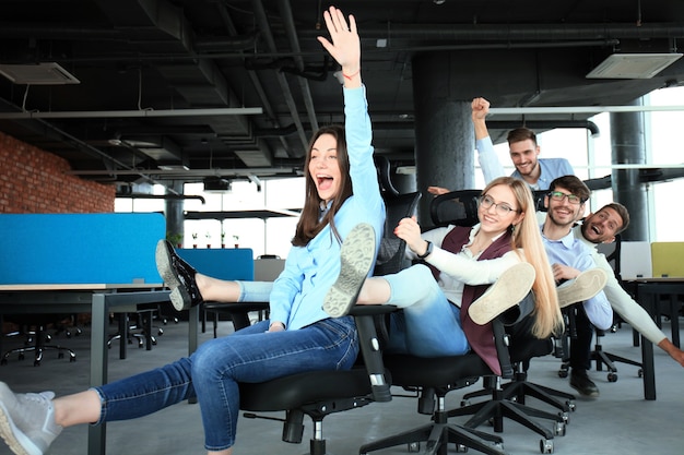 Young cheerful business people in smart casual wear having fun while racing on office chairs and smiling.