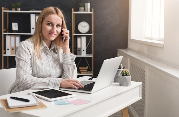 Young cheerful business woman working on laptop and talking on phone at modern office. Business consulting, copy space