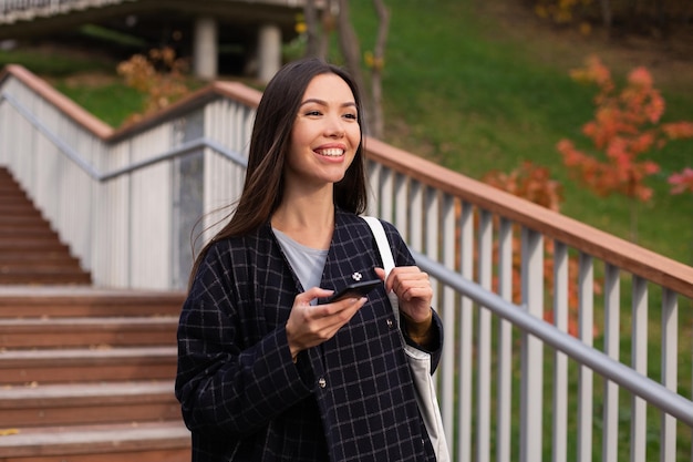 Young cheerful casual woman in coat with cellphone joyfully walking in city park