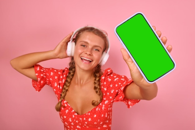 Молодая веселая кавказская женщина в наушниках показывает телефон с зеленым экраном